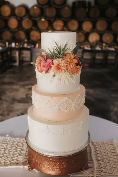 A four-tiered white and peach wedding cake decorated with fresh flowers and intricate piping, displayed on a copper stand.