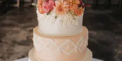 A four-tiered white and peach wedding cake decorated with fresh flowers and intricate piping, displayed on a copper stand.