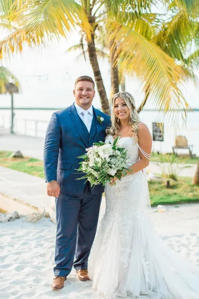 A bride and groom standing on a sandy beach with palm trees in the background.