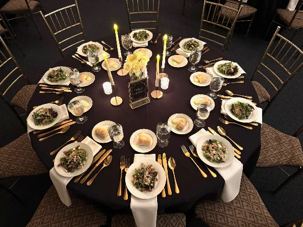 A formally set round dining table with plated meals, gold flatware, and a floral centerpiece.