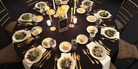 A formally set round dining table with plated meals, gold flatware, and a floral centerpiece.