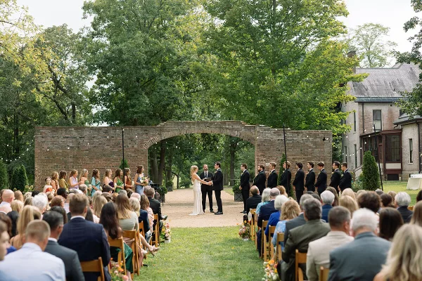 Outdoor wedding ceremony with guests seated on a lawn, featuring a brick archway and a historic Victorian estate in the background.