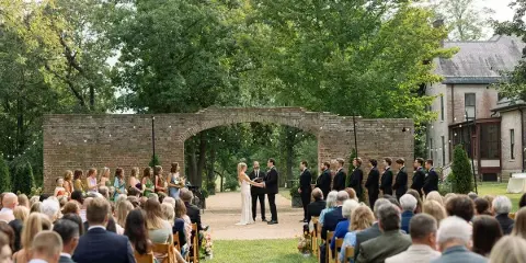 Outdoor wedding ceremony with guests seated on a lawn, featuring a brick archway and a historic Victorian estate in the background.