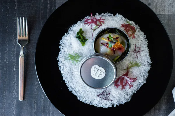 A gourmet plated dish served on a black plate with a fork, featuring a small metal container with a garnished food item and decorative herbs.