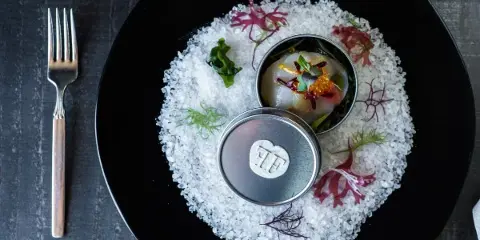 A gourmet plated dish served on a black plate with a fork, featuring a small metal container with a garnished food item and decorative herbs.