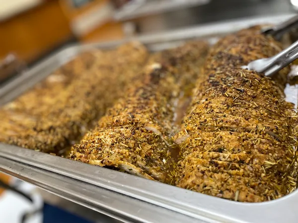 A large, herb-crusted meatloaf sliced and served in a stainless steel catering tray.