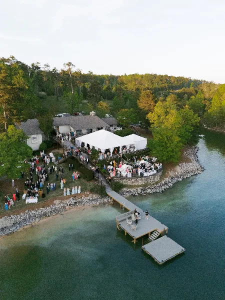 Aerial view of a lakeside wedding reception with white tents and guests gathered on a dock and lawn.