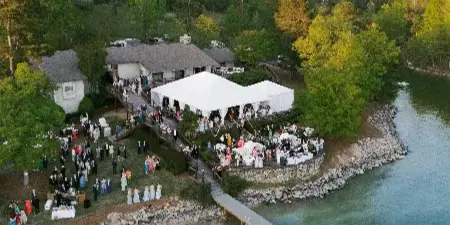 Aerial view of a lakeside wedding reception with white tents and guests gathered on a dock and lawn.
