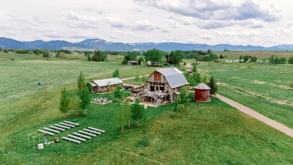 Aerial view of a rustic wedding venue featuring a renovated barn, outdoor ceremony seating, and surrounding green fields with mountains in the background.