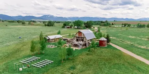 Aerial view of a rustic wedding venue featuring a renovated barn, outdoor ceremony seating, and surrounding green fields with mountains in the background.