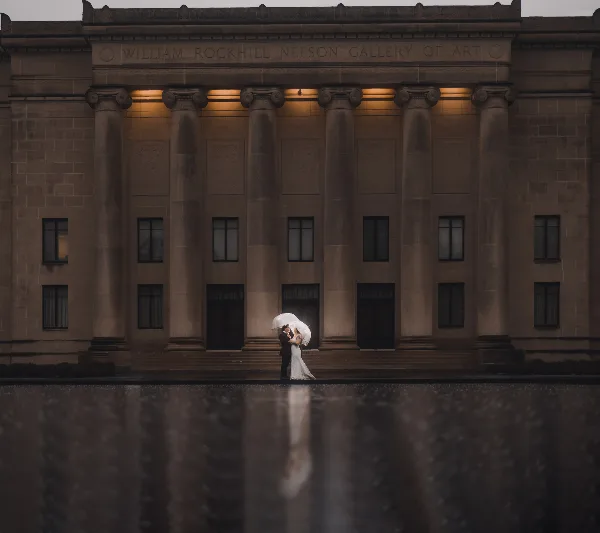 A bride and groom kissing under a white umbrella in front of the William Rockhill Nelson Gallery of Art.
