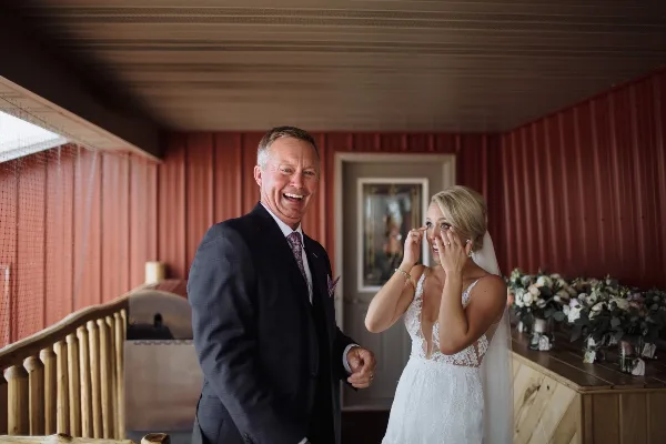 A bride wiping a tear from her eye while standing next to her smiling father in a rustic venue with red corrugated metal walls.