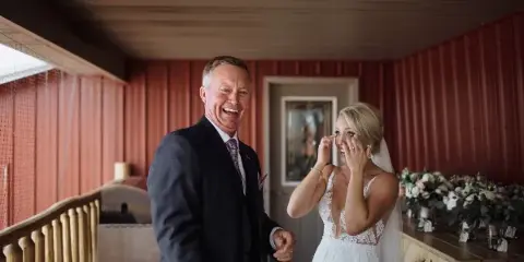 A bride wiping a tear from her eye while standing next to her smiling father in a rustic venue with red corrugated metal walls.