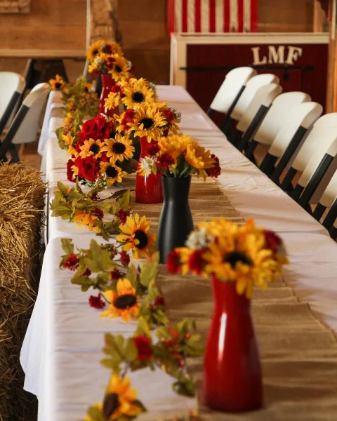 A rustic wedding reception table set with a burlap runner, white chairs, and sunflower centerpieces inside a timber barn.