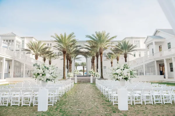 Outdoor wedding ceremony setup with white chairs, floral arrangements, and palm trees at a coastal venue