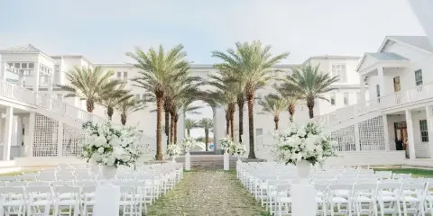 Outdoor wedding ceremony setup with white chairs, floral arrangements, and palm trees at a coastal venue