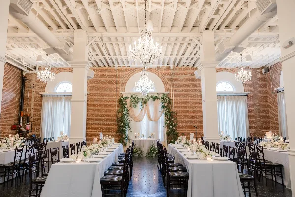 Interior view of a historic brick venue with white columns and chandeliers, set up with long banquet tables for a wedding reception.
