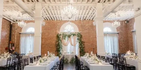 Interior view of a historic brick venue with white columns and chandeliers, set up with long banquet tables for a wedding reception.