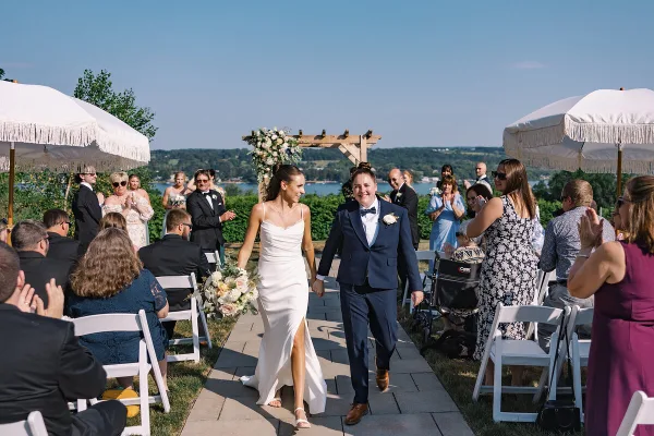 A bride and groom walking down the aisle at an outdoor wedding ceremony with a lake view in the background.