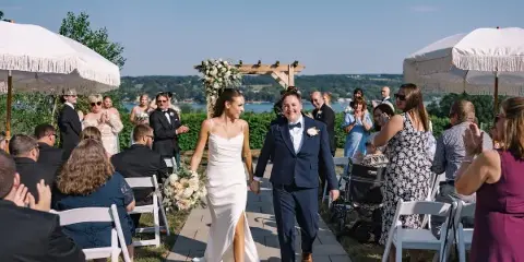 A bride and groom walking down the aisle at an outdoor wedding ceremony with a lake view in the background.