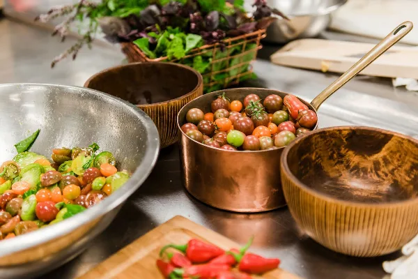 Fresh salad ingredients and heirloom tomatoes prepared in copper bowls and pans on a stainless steel kitchen counter.