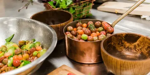 Fresh salad ingredients and heirloom tomatoes prepared in copper bowls and pans on a stainless steel kitchen counter.