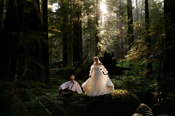 A bride in a white gown and a groom in a white shirt posing in a sunlit redwood forest.