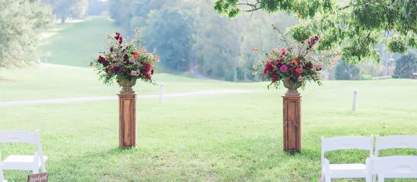 Outdoor wedding ceremony aisle decorated with wooden pedestals and floral arrangements on a grassy lawn.