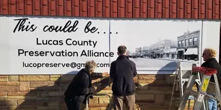 Workers installing a Lucas County Preservation Alliance banner on a historic brick building.