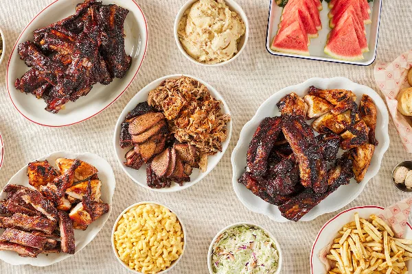 A table spread featuring plates of barbecue ribs, brisket, pulled pork, and chicken alongside bowls of mac and cheese, coleslaw, and potato salad.