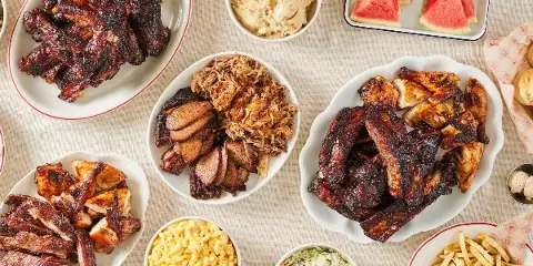 A table spread featuring plates of barbecue ribs, brisket, pulled pork, and chicken alongside bowls of mac and cheese, coleslaw, and potato salad.