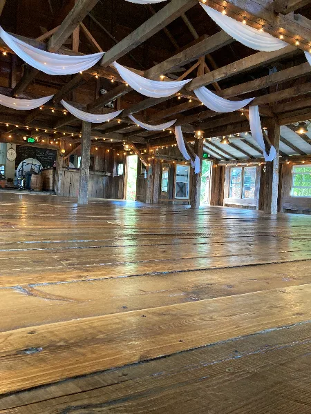 Interior of a rustic wooden barn venue featuring exposed timber beams, string lights, and white fabric draping.