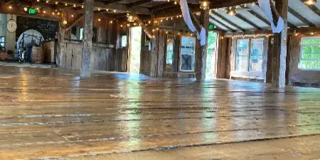 Interior of a rustic wooden barn venue featuring exposed timber beams, string lights, and white fabric draping.