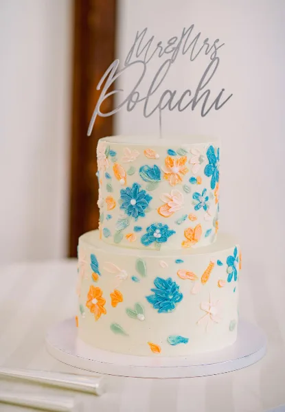 A two-tier white wedding cake decorated with blue, orange, and pink buttercream flowers and a 'Mr & Mrs' topper.
