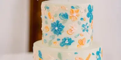A two-tier white wedding cake decorated with blue, orange, and pink buttercream flowers and a 'Mr & Mrs' topper.