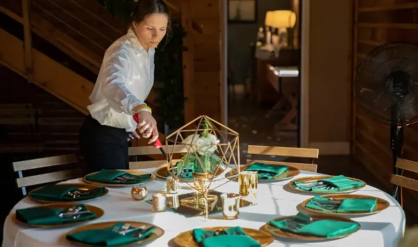 Catering staff member arranging a formal event table with green napkins, gold chargers, and a geometric floral centerpiece.