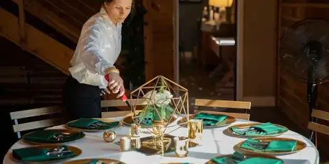Catering staff member arranging a formal event table with green napkins, gold chargers, and a geometric floral centerpiece.