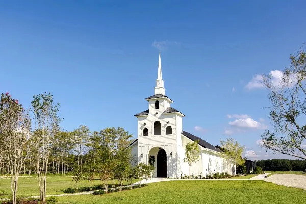 Exterior view of a white chapel with a tall steeple surrounded by green grass and trees under a blue sky.
