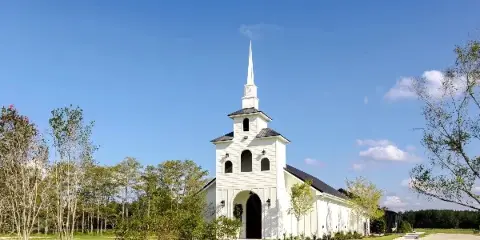 Exterior view of a white chapel with a tall steeple surrounded by green grass and trees under a blue sky.