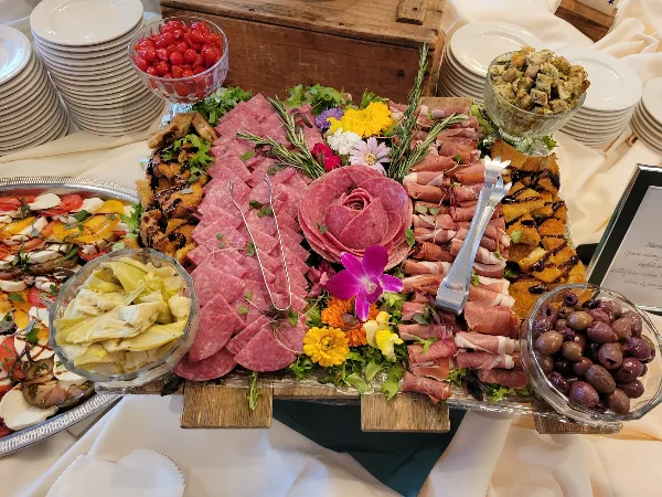 A large, decorative catering platter featuring sliced cured meats, fresh flowers, and bowls of olives and artichokes, set on a table with white plates.