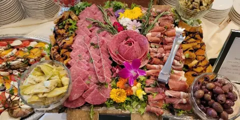 A large, decorative catering platter featuring sliced cured meats, fresh flowers, and bowls of olives and artichokes, set on a table with white plates.