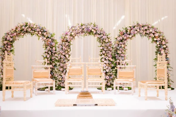 A white wedding stage featuring three floral arches, traditional chairs, and a ceremonial altar setup.