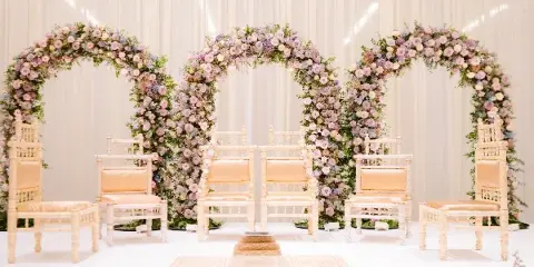 A white wedding stage featuring three floral arches, traditional chairs, and a ceremonial altar setup.