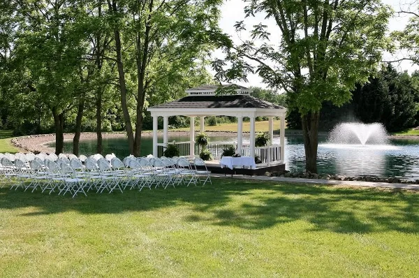 Outdoor wedding ceremony setup with white chairs arranged in front of a white gazebo near a pond with a fountain.