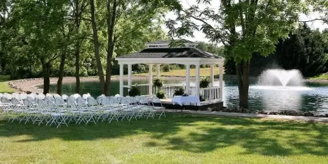 Outdoor wedding ceremony setup with white chairs arranged in front of a white gazebo near a pond with a fountain.