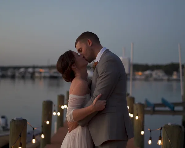 A bride and groom kissing at a waterfront venue during twilight with string lights in the foreground.