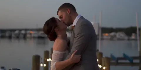 A bride and groom kissing at a waterfront venue during twilight with string lights in the foreground.