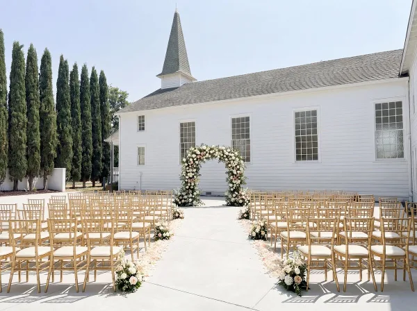 Outdoor wedding ceremony setup with floral arch and chairs in front of a white historic chapel building.