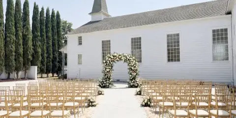 Outdoor wedding ceremony setup with floral arch and chairs in front of a white historic chapel building.