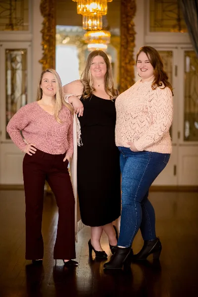 Three smiling women standing together in a formal interior with chandeliers and gold-framed mirrors.